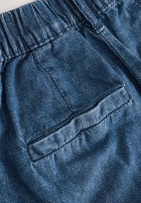 Mėlynas džinsinis audinys su švelnia tekstūra, turintis nugaros kišenę ir elastinę juosmenį. Paviršiuje matyti subtilūs spalvų niuansai.