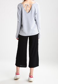 Maglietta grigia chiara a maniche lunghe con scollatura a V profonda e dettaglio con laccio, abbinata a pantaloni neri a gamba larga e sneaker bianche con accenti rossi.