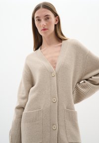 Beige stickad cardigan med V-ringning, knappar fram och ribbade ärmslut. Har två framfickor med ett strukturerat mönster.