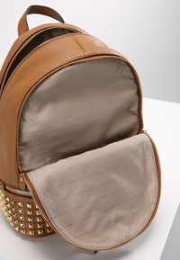 Pieni ruskea nahkareppu, jossa on kultaiset niitit, avoin pääosasto ja vaaleanbeige kangaspinnoite, jossa on toistuva brändinimi.