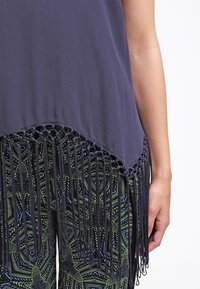 Personne portant un haut violet foncé à ourlet à franges sur un pantalon à motifs géométriques verts et bleus, bras détendu le long du corps.