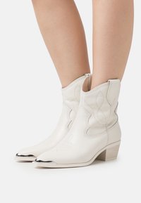 Botas de vaquero de cuero blanco con costuras decorativas, puntera puntiaguda y tapa de punta con acentos metálicos. Presentan un tacón bajo y un diseño con recortes.
