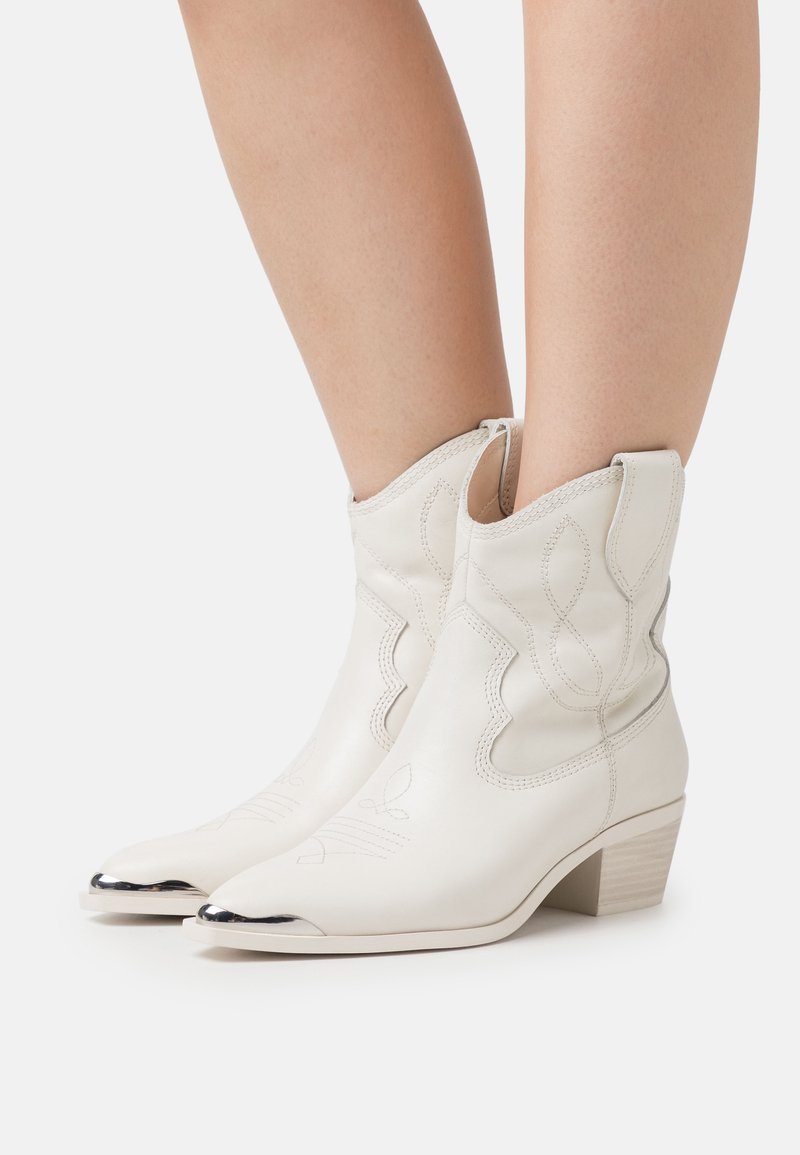 Botas de vaquero de cuero blanco con costuras decorativas, puntera puntiaguda y tapa de punta con acentos metálicos. Presentan un tacón bajo y un diseño con recortes.