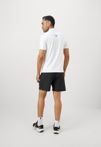 Homem de costas vestido com uma camisola pólo branca com um logótipo nas costas, calções pretos, meias brancas e sapatilhas de desporto pretas.