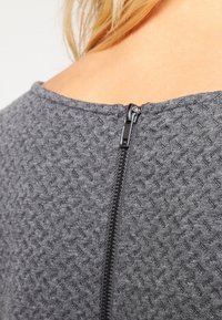 Tissu gris texturé avec un motif géométrique. Dispose d'une fermeture à glissière noire à l'arrière, ajoutant un détail au design du vêtement.