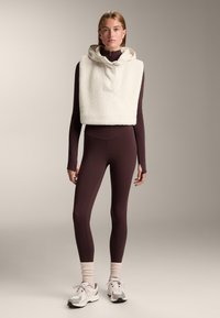 Ivoorkleurige fleece cropped hoodie met hoge kraag, gedragen over een donkerburgundy langemouwtop en bijpassende legging. Voorzien van ribgebreide manchetten en witte sneakers.