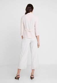 Ljusrosa jacka med trekvartslånga ärmar och en baktillgång, i kombination med vita randiga vida byxor och svarta klackskor.