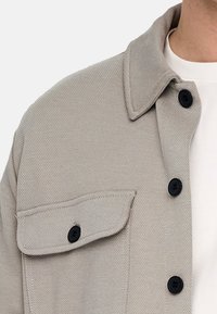 Hellbeige Jacke mit schwarzen Knöpfen, Klappentasche auf der Brust, getragen über einem weißen Rundhals-T-Shirt.