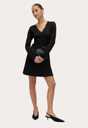 Mujer con un vestido negro de manga larga y escote en V con borde de encaje, sosteniendo un bolso negro y llevando tacones negros con tira trasera.