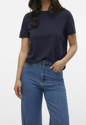 Frau trägt ein marineblaues Kurzarm-T-Shirt, das in hoch taillierte blaue Jeans gesteckt ist, mit einer Hand in der Tasche.