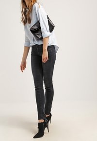 Camisa de botonadura azul claro con mangas remangadas, combinada con jeans ajustados oscuros. Tacones negros de punta con recortes en el tobillo y un bolso de mano negro.