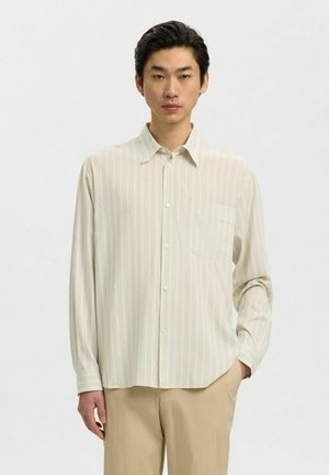Jeune homme portant une chemise beige clair rayée avec boutons, une main dans la poche, debout devant un fond blanc uni.