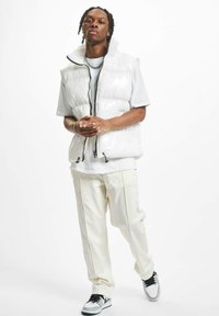 White puffer vest with high collar, glossy texture, front zipper, worn over a light grey t-shirt and paired with cream pants and black sneakers.