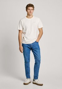 Cream cotton t-shirt with short sleeves, paired with blue denim jeans. Model wearing white sneakers with beige accents. Simple, casual look.