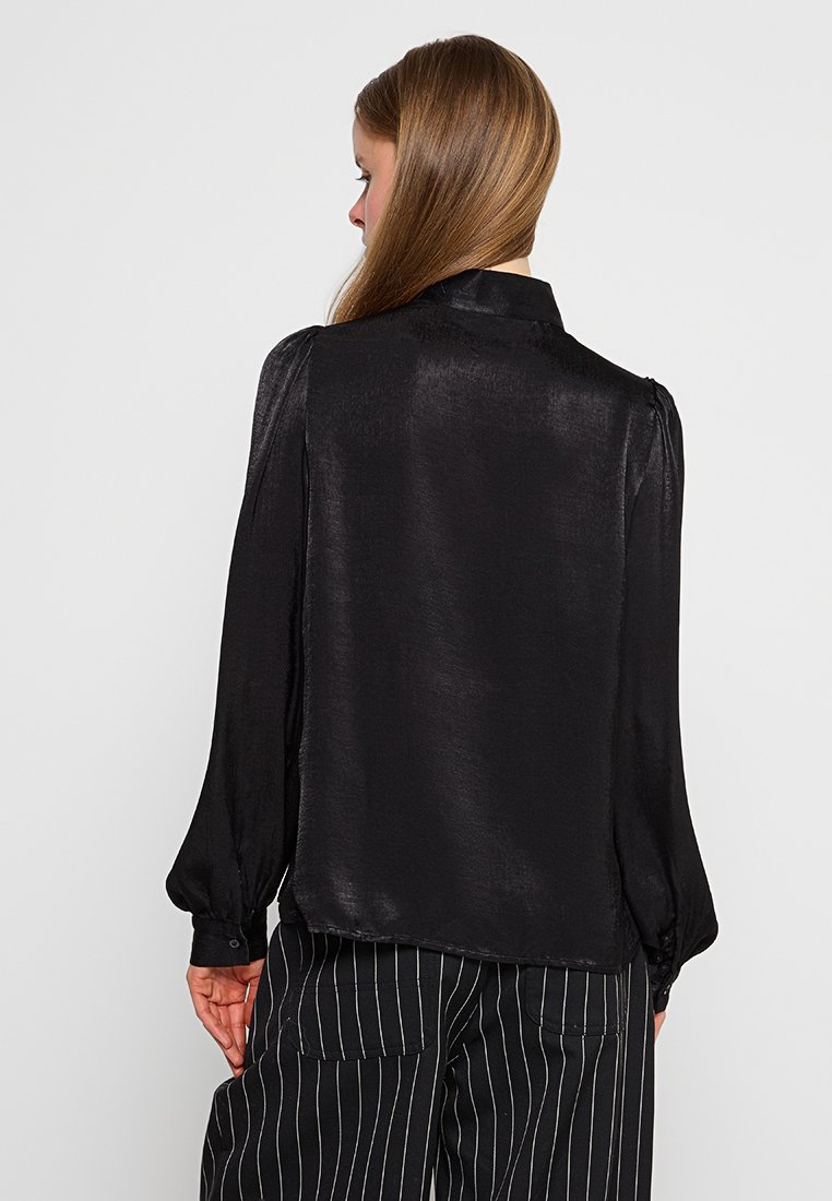 Blusa de seda negra con mangas largas y fruncidas, un cuello clásico y una textura ligeramente translúcida. Combinada con pantalones a rayas en blanco y negro.