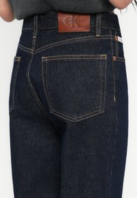 Blugi din denim albastru închis cu un finisaj texturat, având cusături portocalii și o etichetă maro din piele cu logo-ul Calvin Klein pe talie.