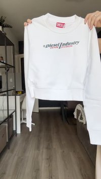 Bela skraćena dukserica od pamuka s rebrastim manžetama. Na prednjoj strani se nalazi logo "Diesel Industry Denim Division" u sivoj i crvenoj boji.