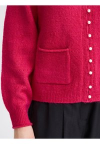 Spilgti rozā adīta jaciņa ar teksturētu rakstu, pogu aizdare ar pērļu līdzīgām pogām un viena priekšējā kabata.