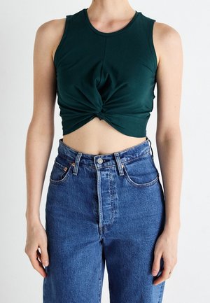 Persoon draagt een donker groen mouwloos crop top met een gedraaide knoop aan de voorkant, gecombineerd met een high-waisted blauwe spijkerbroek.