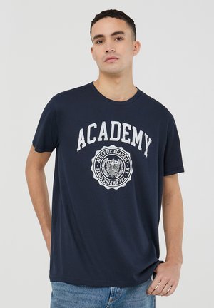 T-shirt in cotone blu navy con maniche corte, con la scritta bianca "ACADEMY" e un logo grafico circolare sul davanti. Vestibilità casual.