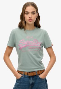 Camiseta de algodón verde claro de manga corta, con un diseño gráfico de logo en rosa y texto. Combinada con un pantalón de mezclilla de cintura alta y un cinturón marrón.