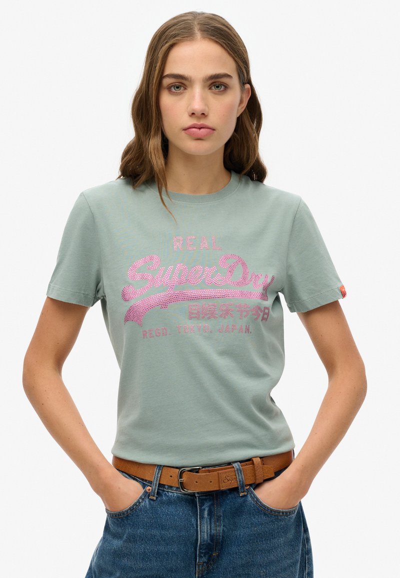 Camiseta de algodón verde claro de manga corta, con un diseño gráfico de logo en rosa y texto. Combinada con un pantalón de mezclilla de cintura alta y un cinturón marrón.