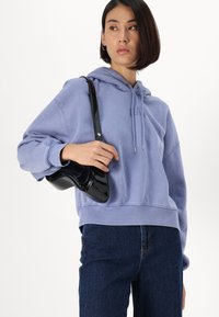 Kvinne iført en lys lilla hettegenser og mørke jeans, som holder en liten skinnende svart skulderveske, og ser mot høyre.