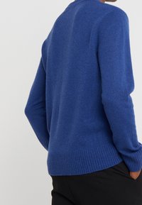 Niebieski sweter dziergany o fakturowanym wykończeniu, z prążkowanym dołem i okrągłym dekoltem, widziany od tyłu, w połączeniu z czarnymi spodniami.