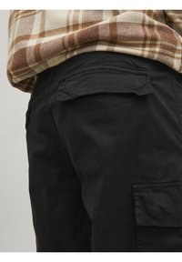 Pantalon cargo noir avec une poche latérale à rabat et une texture lisse. Une chemise à carreaux marron et crème émerge partiellement au-dessus.