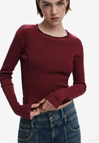 Burgunderfarbenes, geripptes Langarmshirt mit Rundhalsausschnitt und rohen Kanten, kombiniert mit hoch-taillierten blauen Denim-Jeans.