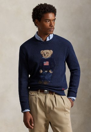 Homme portant un pull bleu marine avec un ours en peluche portant un pull à motif de drapeau, une chemise bleu clair à col et un pantalon beige plissé, mains dans les poches.