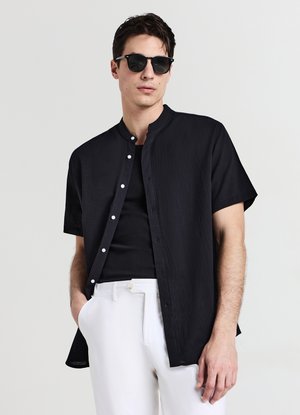 Chemise noire à manches courtes avec col droit, tissu texturé et boutons blancs, associée à un débardeur noir et un pantalon blanc.