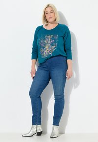 Tyrkysová mikina s grafickým motivem "MAGIC", spárovaná s úzkými modrými džíny a stříbrnými kotníkovými botami. Měkká látka, uvolněný střih.