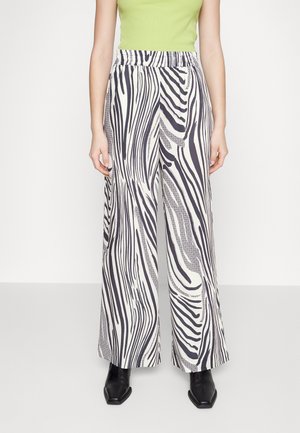 Femme portant un pantalon large taille haute à imprimé zèbre noir et blanc, associé à un haut sans manches vert citron et des chaussures noires à bouts pointus.