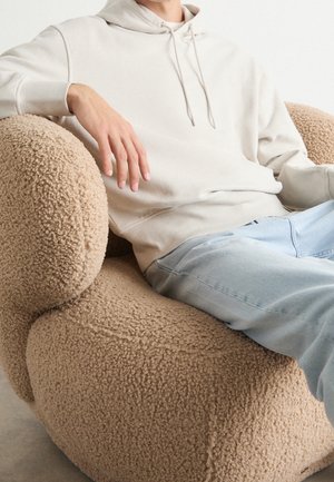 Person i beige hættetrøje og lyse jeans, der sidder i en rundet, tekstureret beige lænestol med blødt, krøllet stofbetræk.