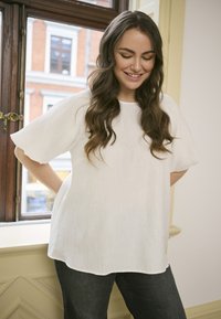 Blusa bianca a maniche corte e tessuto strutturato con vestibilità ampia. Dettaglio di pieghe notevole e orlo curvo, abbinata a jeans scuri.