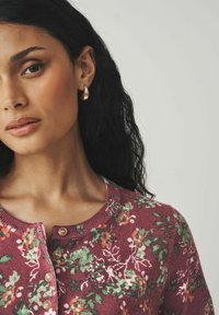 Femme avec des cheveux bouclés foncés portant un pull à boutons floral bordeaux et des boucles d'oreilles en forme de goutte dorées sur un fond uni.
