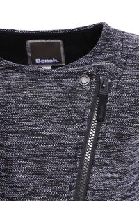 Czarna i szara teksturowana kurtka materiałowa z zamkiem błyskawicznym oraz okrągłym metalowym detalem logo przy kołnierzu, zaprojektowana w dopasowanym kroju.