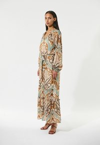 Robe longue à motifs dans des tons chauds de marron, turquoise et crème. Elle présente des manches longues, une ceinture et un ourlet à volants. Portée avec des sandales marron.