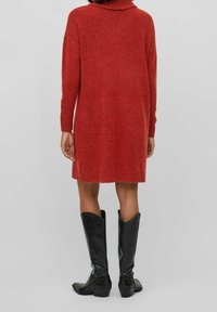 Robe-pull rouge en maille avec un col montant, des manches longues et une coupe décontractée. Associée à des bottes noires montantes à talon léger.
