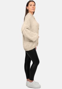 Cardigan oversize beige lavorato a maglia con grandi bottoni, tessuto testurizzato e spalle basse, abbinato a leggings neri e sneakers bianche.