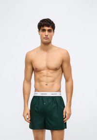 Bărbat fără cămașă, cu păr scurt și închis la culoare, purtând șorturi boxer Lacoste de culoare verde închis, cu talie albă, stând în fața unui fundal alb simplu.