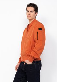 Orangene Bomberjacke mit Reißverschlussfront, gerippten Bündchen und Saum. Verfügt über eine mit Reißverschluss gesicherte Tasche am linken Ärmel und ein Logo-Patch.