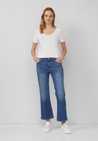 Witte V-hals T-shirt, gecombineerd met blauwe wijde jeans, met een knoopsluiting en voorzakken, gedragen met witte sneakers op een grijze achtergrond.
