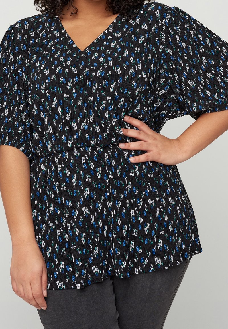 Femme portant un chemisier noir avec de petites fleurs blanches et bleues, un col en V et des manches bouffantes, associé à un pantalon gris foncé.