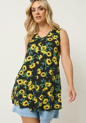 Blondynka w czarnej bezrękawnikowej tunice z żółtym nadrukiem słonecznika i jasnoniebieskich jeansowych szortach, stojąca na jednolitym tle.