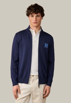 Junger Mann trägt navyblauen Reißverschluss-Cardigan mit blauem "H"-Patch über weißem Polo und beiger Hose, stehendes vor neutralem Hintergrund.