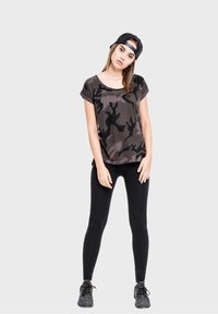 Camo-print short-sleeve t-shirt in dark tones, paired with black leggings and patterned sneakers. Model wears a black cap.