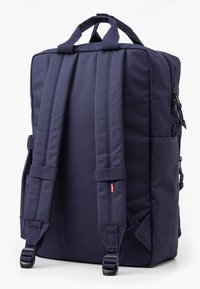 Tamsiai mėlynas kuprinės krepšys su paminkštintomis petnešomis ir viršutine rankena, pasižymintis struktūruotu dizainu ir šonine kišene daiktams laikyti.