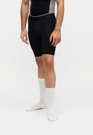 Mand iført sorte kompressionsshorts, en tætsiddende atletisk top og lange hvide sokker stående på en lys baggrund.